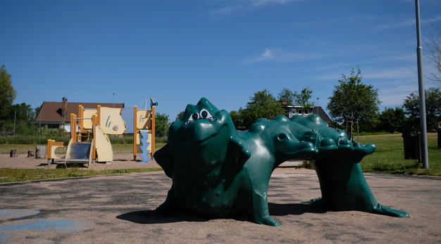 Fotografiet är ett foto på ett klätterredskap som ser ut som en drake. Redskapet är stort och grönt. Drakens ansikte är glatt och har ett stort leende. I bakgrunden syns en lekställning, flera hus och en blå himmel. 