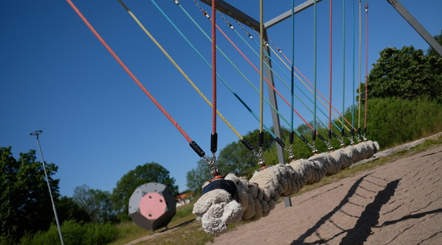 Bilden föreställer en balansgångställning som är en bit upp i luften. Delen man går på är gjord i rep och är ljust beige. Från gångdelen är de rep man kan hålla i. Repen är i olika färger. Det finns blåa, rosa, gula och gröna. Marken under är sand. 