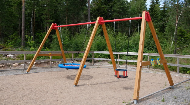 Bilden föreställer en gungställning i trä med två platser. Bilden är tagen snett från sidan. Gungan längst bort är en blå kompisgunga och gungan närmast i bild är en gunga för små barn. I bakgrunden syns mycket grönska och även ett staket i trä som omringar gungorna. 