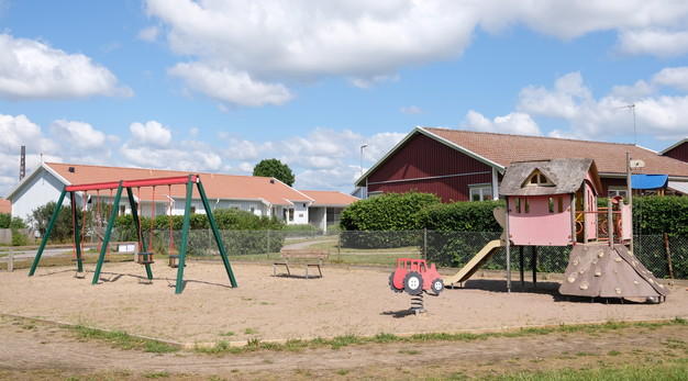 Fotografiet är en översiktsbild över Linängens lekplats. Från vänster i bild syns en lekställning, ett gungdjur i form av en traktor och en stor röd och gul lekställning med rutschkana. Marken är sand och i bakgrunden syns två stora hus. Himlen är blå. 