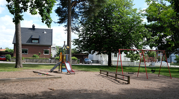 Fotografiet är en bild över Lövdungens lekplats. Till vänster i bild finns en liten lekställning med en röd rutschkana och en mindre klättervägg. Till höger i bild finns en lekställning med två gungor. Lekställningen är röd. I bakgrunden syns två bostadshus.