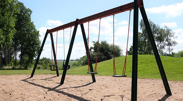 Bilden föreställer en gungställning med fyra gungor. Ställningen är grön och detaljerna på gungorna är röda. I bakgrunden syns en grön kulle. Marken under gungorna är sand. Solen skiner på bilden och gör att det blir mycket värme i den. 