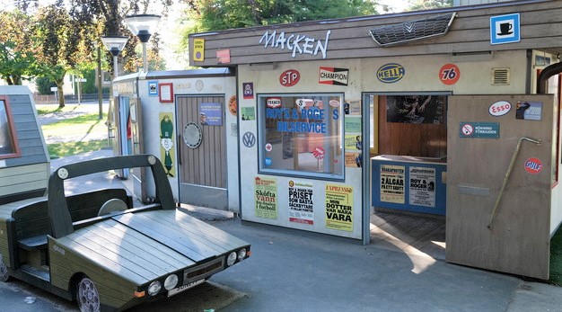 Bilden föreställer ingången till macken-verkstaden på lekplatsen Macken. På verkstaden sitter alla möjliga typer av klistermärken och även löpsedlar från olika tidningar. Klistermärkena är bland annat en wolkswagen logotyp, en skylt med en kaffekopp och en skylt där det står Champion. Framför ingången till verkstaden står det en grön träbil. 