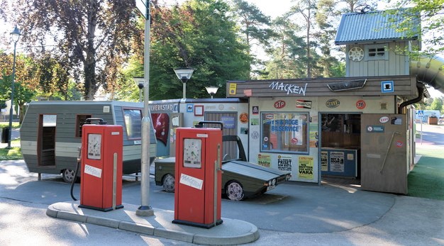 Bilden är ett fotografi över bensinmacken och verkstaden på Macken-lekplasten. I förgrunden står två röda pumpstationer. I mitten av dessa är en grå lyktstolpe. Bakom bensinpumparna står en grön bil och husvagn parkerade. Dessa är byggda i trä. Bakom dessa syns ingången till macken-verkstaden. Till höger i bild uppe på verkstaden finns ett grått litet hus. 