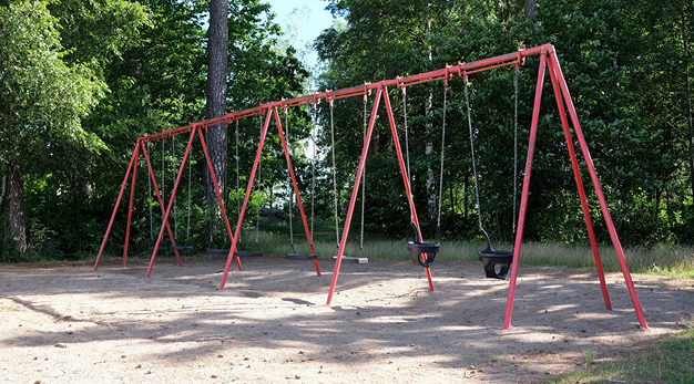 Foto är en bild på en röd gungställning med åtta gungor. Fotografiet är taget snett framifrån och de två gungorna närmast i bild är gungor för småbarn. Marken under är sand och det ligger kottar över hela marken. I bakgrunden är det tät skog.