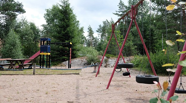 Bilden är en översiktsbild över lekplatsen Ravinen. Till höger i bild står en gungställning. Den är fotograferad från sidan. Gungställningen är röd och har fyra gungor. Till vänster i bild syns en lekställning i grönt, gult, blått och rött. Den har en rutschkana och ett stort klätternät. I bakgrunden syns ett litet berg och flertalet träd. 