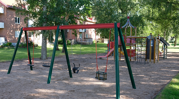 I bildens förgrund står en grön och röd gungställning med fyra gungor. I bakgrunden står en stor lekställning med rutschkana, klätternät och ett litet hus. Marken är sand och över lekplatsen är det grenar från träd som hänger ner. Bilden förmedlar en somrig känsla. 