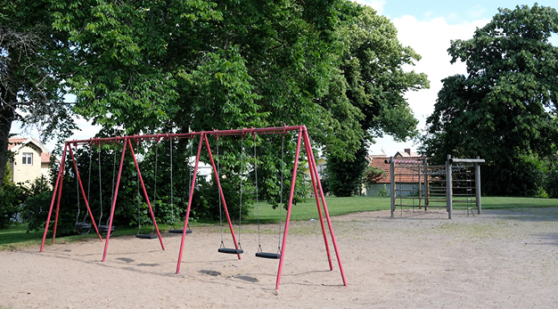 Bilden är en översiktsbild över Sandhagens lekplats. Till vänster står en gungställning med sex stycken gungor. Till höger i bild syns en jättestor fyrkantig klätterställning. I bakgrunden syns flera stora gröna träd. 
