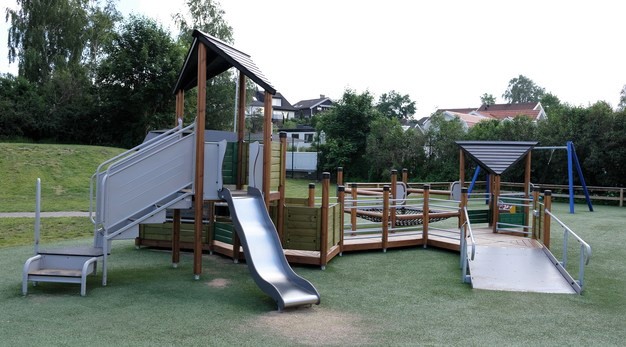 Bilden föreställer en stor lekställning bestående av ett hus och och flera gångar i marknivå. Lekställningen är anpassad för rullstol så det finns en ramp till höger. Huset har en rutschkana. Marken är konstgräs och i bakgrunden syns en blå gungställning. 