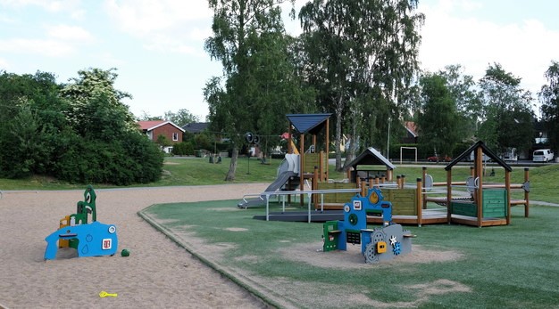 Fotografiet är ett foto av en lekställning och två små lekstationer. Till vänster i bild är den första lekstationen, den är blå, gul och grön och har olika små fönster. Marken under lekstationen är sand och där ligger en gul spade. I bakgrunden finns en stor lekställning med rutschkana. Framför ställningen finns den andra lekstationen. Den är grön, blå och grå och har muttrar på sig. Marken under denna och lekställningen är konstgräs.  