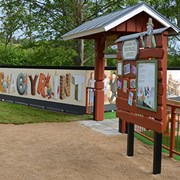Bilden föreställer entrén till Skrotnisses lekplats. I entrén finns en stor skylt som hälsar besökare välkomna till lekplatsen. I bild syns en parkbänk och ingången samt utsidan av den labyrint som finns på lekplatsen.  