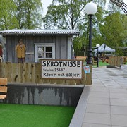 Bilden är en vybild över en del av lekplatsen. Rakt fram syns en grå bod och en stor skylt där det står "Skrotnisse, köper och säljer".  I förgrunden står en bänk i ljust träslag. 