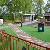 Bilden föreställer Skrotnisses lekplats innan invigningen. Till vänster i bild finns ingången till en labyrint. I bild syns även två små hus i grått och i rött. Marken är delvis täckt med grön markduk och delvis täckt med vad som ska efterlikna ett tågräls. I bakgrunden syns stora gröna björkar.  