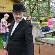 Bilden föreställer en man i svart kostym och hatt. Han står framför barn som leker