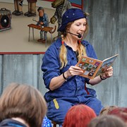 Bilden föreställer en kvinna som sitter på en pall och läser ur en bok. Kvinnan är klädd i en blå overall och en blå keps som hon har bak och fram. Kvinnan ser väldigt glad och entusiastisk ut. I förgrunden syns personer som står och lyssnar på sagostunden.  