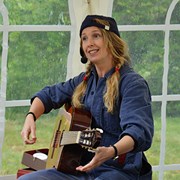 Bilden föreställer en kvinna som sjunger och spelar gitarr. Hon är klädd i blå keps och blå overall. 