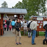 Bilden föreställer entrén till Skrotnisses lekplats under invigningen av lekplatsen. Det är massor med barn och vuxna vid entrén och inne i lekparken. Två personer i förgrunden håller i paraplyn och flera personer bär regnkläder. Trots att regnet ligger i luften är det många leenden i parken. 