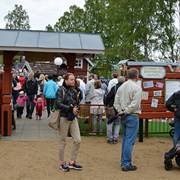 Bilden föreställer entrén till Skrotnisses lekplats under invigningen av lekplatsen. Det är massor med barn och vuxna vid entrén och inne i lekparken. Två personer i förgrunden håller i paraplyn och flera personer bär regnkläder. Trots att regnet ligger i luften är det många leenden i parken. 