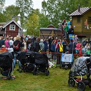 Bilden är tagen utanför lekplatsen på invigningsdagen. I bild syns vuxna och barn som utforskar stadens nya lekplats. I bakgrunden syns barn som köar för att få åka rutschkana. I förgrunden står flera vuxna och småpratar. 