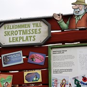 Bilden föreställer den tavla som man som besökare möts av vid entrén till Skrotnisses lekplats. På tavlan, som är röd, står det med stora bokstäver "Välkommen till Skrotnisses lekplats". 