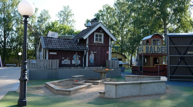 Bilden är ett fotografi från Skrotnisses lekplats. I förgrunden finns en sandlåda formad som en hexagon. I bakgrunden står en stuga i rött. På stugans fasad sitter karaktärer från böckerna om skrotnisse. Bredvid stugan står en båt i gult och rött. På denna står det Balder. 