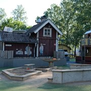 Bilden är ett fotografi från Skrotnisses lekplats. I förgrunden finns en sandlåda formad som en hexagon. I bakgrunden står en stuga i rött. På stugans fasad sitter karaktärer från böckerna om skrotnisse. Bredvid stugan står en båt i gult och rött. På denna står det Balder. 