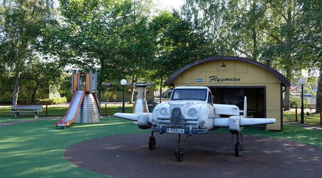 Bilden föreställer ett flygplan i grått. Flygplanet står på en rund helikopterplatta. I bakgrunden syns en gul hangar. Taket är rundat och det står Flygmotor på det. I bakgrunden syns en rutschkana och en grå parkbänk. 