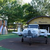 Bilden föreställer ett flygplan i grått. Flygplanet står på en rund helikopterplatta. I bakgrunden syns en gul hangar. Taket är rundat och det står Flygmotor på det. I bakgrunden syns en rutschkana och en grå parkbänk. 