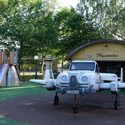 Bilden föreställer ett flygplan i grått. Flygplanet står på en rund helikopterplatta. I bakgrunden syns en gul hangar. Taket är rundat och det står Flygmotor på det. I bakgrunden syns en rutschkana och en grå parkbänk. 