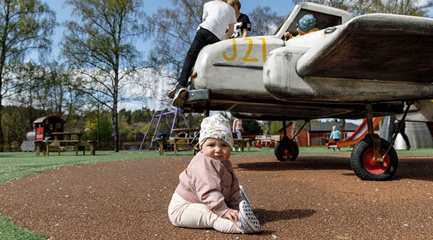 Bebis som sitter på backen i lekplats, flygplan med lekande barn i bakgrunden. 