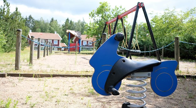 Bilden är en närbild på ett blått gungdjur formad som en motorcykel. I bakgrunden syns ett staket som omringar en gungställning. Fotografiet är taget i grodperspektiv och marken är sand. 