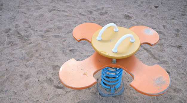 Bilden föreställer ett gungdjur med fyra platser. Sittdelen är rund och orange. Marken under är sand. Den orangea färgen lyser i bild. 