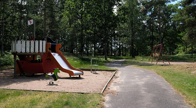 Fotografiet är en översiktsbild över en lekplats. I mitten går en asfalterad väg. Till vänster i bild syns en lekställning i form av en båt och till höger om vägen finns en gungställning i rött. 