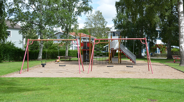 Bilden är en översiktsbild över lekplatsen Solgläntan. I förgrunden står två stora röda gungställningar och bakom dem syns en lekställning med rutschkana. Marken i lekplatse är sand och runt omkring är det gräsmatta. 