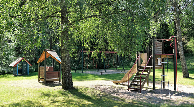 Bilden är ett översikts foto över spelmansparkens lekplats. Mitt i bild står en stor björk. Till höger om den finns en stor lekställning med en gul rutschkana. Till vänster om björken finns två små lekhus. Den ena lekhuset är blått med rött tak och det andra huset är rött med gult tak.