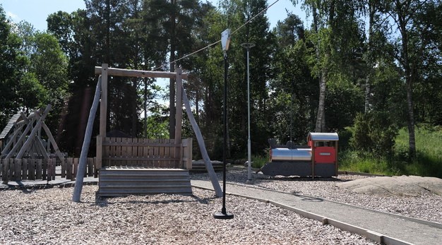 Bilden föreställer en linbana. I bild ser man avsatsen och sittdelen. Marken under är träflis och i bakgrunden syns ett lekhus i form av ett tåg och en skogsdunge. 