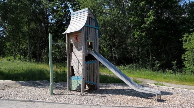 Bilden föreställer en lekställning med rutschkana och ett klätternät. Ställningen är smal och hög och är skapad i trä. Marken är träflis och i bakgrunden syns tät skog. 