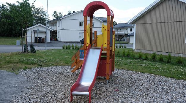 Bilden föreställer en liten lekställning med rutschkana. Ställningen är röd, gul och träfärgad. Marken under är träflis och i bakgrunden syns ett stort vitt hus och husknuten på ett beige. 