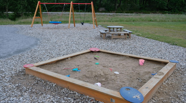 Bilden föreställer en fyrkantig sandlåda. I sandlådan är det massa olika redskap för sandlek. I bakgrunden syns ett parkbord och en gungställning med en vanlig gunga och en kompisgunga. 