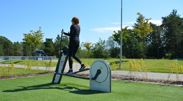 Bilden föreställer en kvinna klädd i svarta träningskläder och uppsatt hår som tränar på en crosstrainer utomhus. Marken under är konstgräs och i bakgrunden syns en grusstig och en stor gräsmatta. 