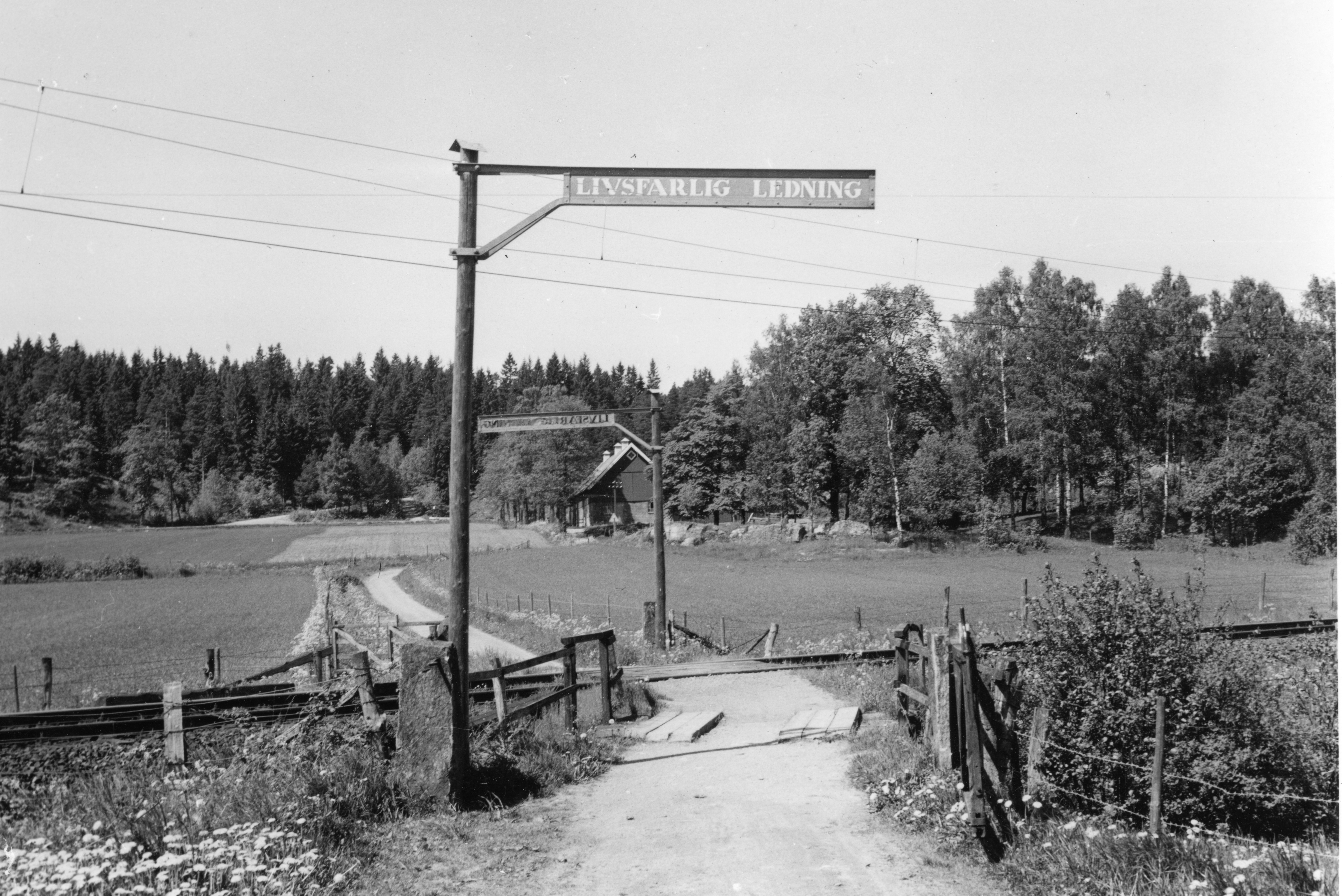 Svartvit bild på en gammal järnväg. På bilden syns en skylt med texten: Livsfarlig ledning.