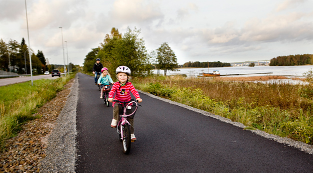 Två barn och en vuxen kommer cyklandes utmed en rak och nyasfalterad cykelväg