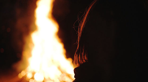 Närbild på en eld. En kvinna står i förgrunden och tittar in i elden.