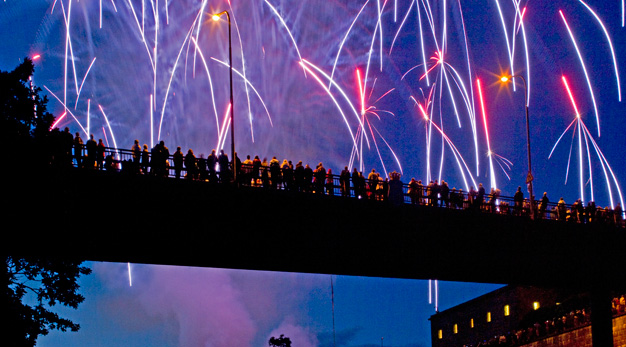 Människor på en bro tittar upp mot fyrverkerierna mot en blålila himmel