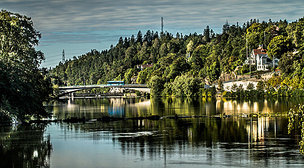 Fotot visar en vy över kanalen i Trollhättan. Vattnet är spegelblankt och det är prunkande grönska utmed vattnet I bakgrunden ser man Spiköbron och en buss som kör över den.