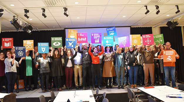 Politiker, tjänstemän och representanter för de kommunala bolagen i Trollhättan hade samlats för att diskutera hur Trollhättan kan bidra i genomförandet av Agenda 2030 – och dess 17 globala mål för en hållbar utveckling. Personerna håller upp skyltar för alla de 17 målen.