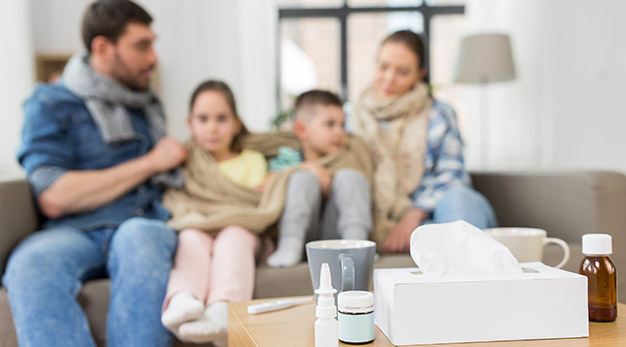 Medicin på bordet i förgrunden, och sjuk familj i hemmet i bakgrunden