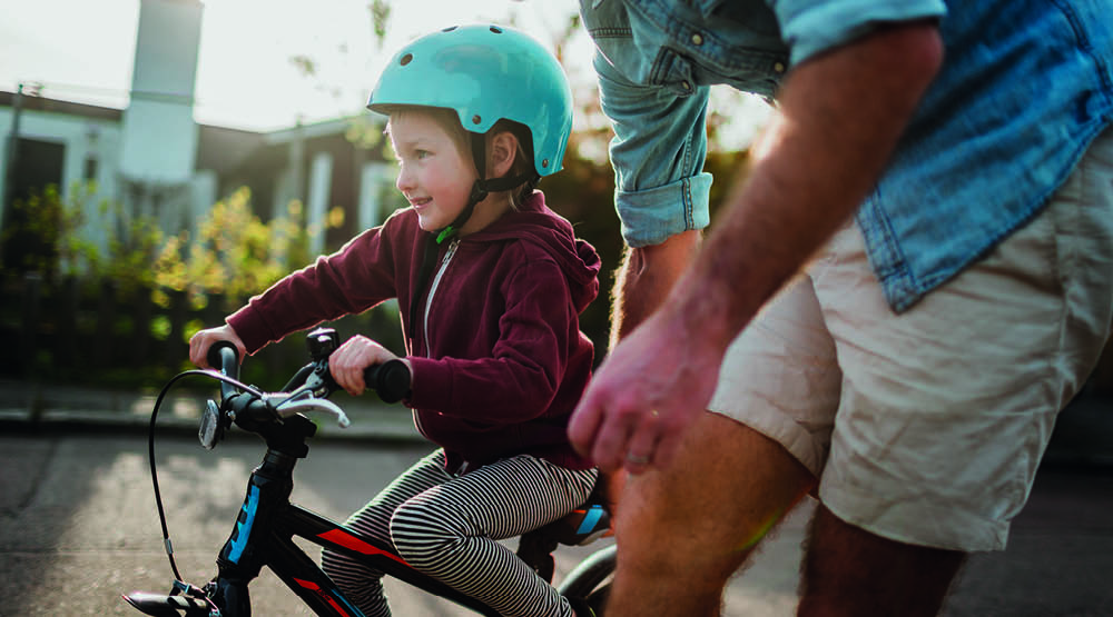 Bild på barn med cykelhjälm som sitter på en cykel. En man lutar sig över och håller i cykeln.