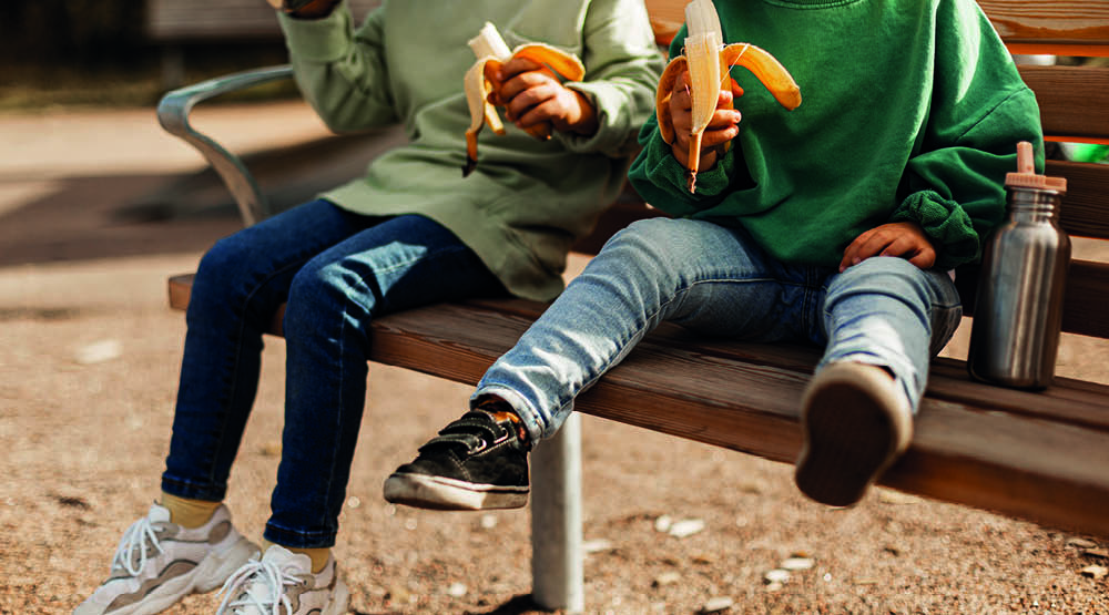 Bild på fyra barnsben som sitter på en parkbänk och äter banan. Bilden är beskuren vid axlarna på barnen.