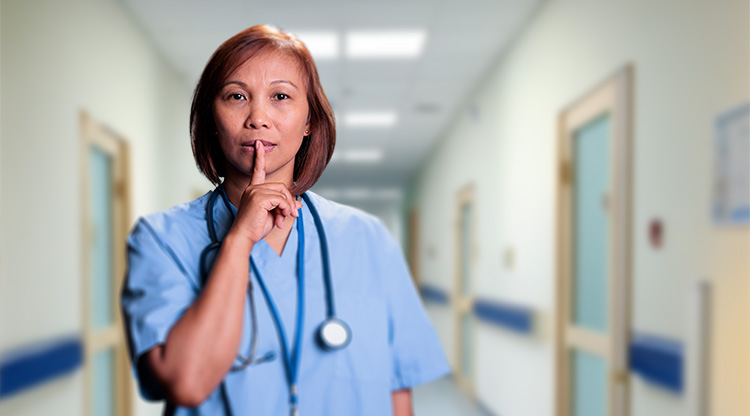 En kvinnlig läkare står i en sjukhuskorridor med ett finger pekandes framför munnen.